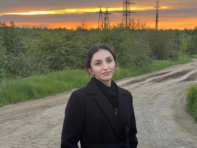 LeonieRahim with small tits, wearing a black coat, standing on a gravel road, green bushes, and power lines at sunset, smiling, hands by sides, orange sky in background.