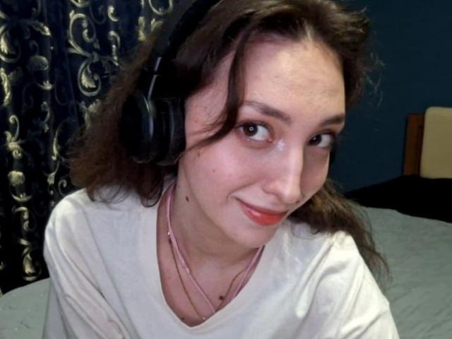 LeonieRahim with medium brown wavy hair, wearing black headphones, a white shirt, and pink necklace, smiling subtly, red lipstick, blue wall, and dark floral curtain in the background.
