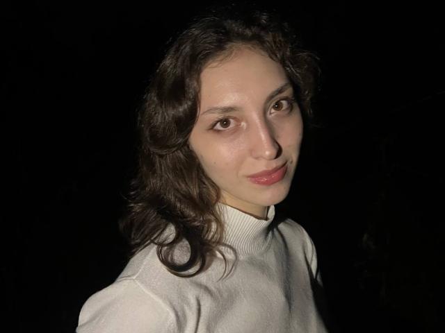 LeonieRahim with wavy dark brown hair, pale skin, and brown eyes, wearing a white turtleneck, smiling slightly, dark background, visible light source from the left.