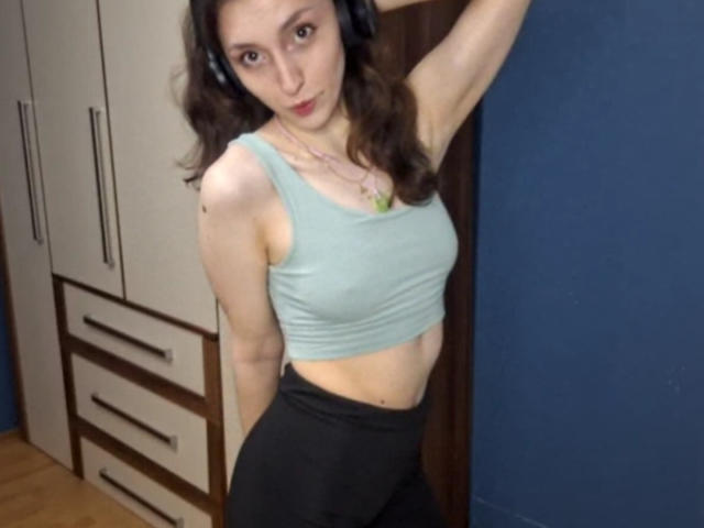 LeonieRahim with small tits, wearing a light green tank top and black pants, standing in a room, arm raised, headphones on, looking at the camera.