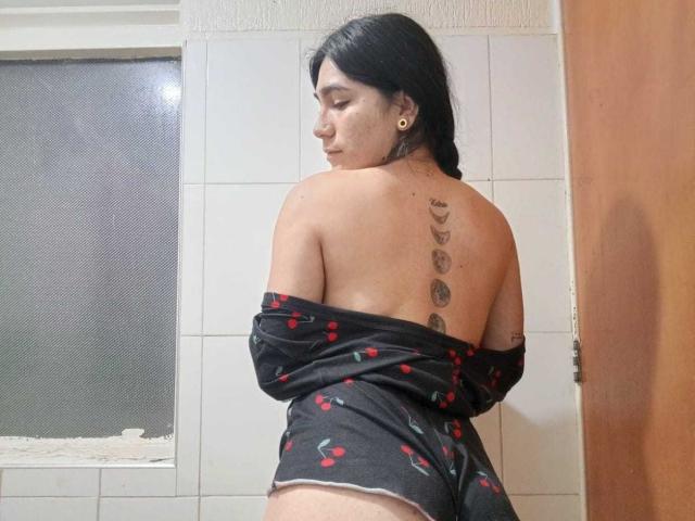 ZoeeRoux with bare back, black shirt off-shoulder, cherry print, tattoo line, medium brown skin, black hair, small gold earrings, standing in bathroom, looking back, smiling.