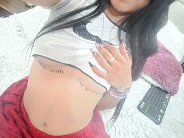 ZoeeRoux with long black hair, white crop top, red plaid shorts, visible tattoos on her ribcage, and a ring on her finger, holding her shirt.