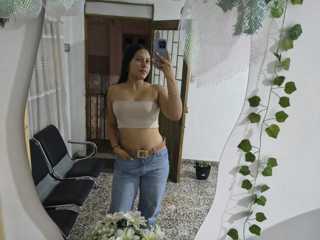 MarilynHills with long black hair, tan skin, small tits in a beige strapless top, blue jeans, and a brown belt, taking a mirror selfie, hands in pockets.