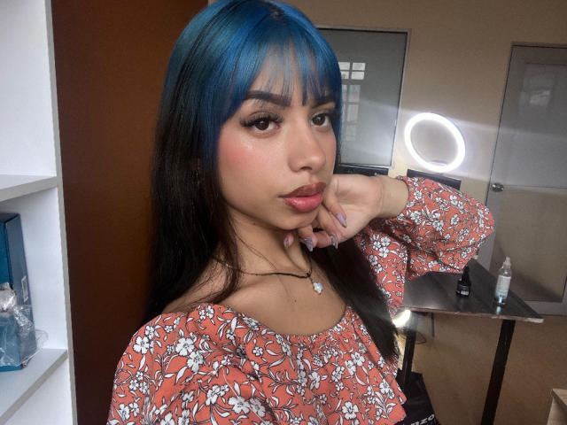 Kazumii with blue-black hair, floral dress, and a necklace, has her finger on her cheek, showing her pink lips and shiny skin. Ring light in background.