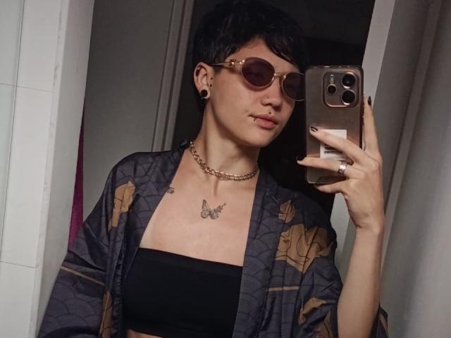 LissyLuxe with short black hair, wearing round sunglasses, a black tube top, floral kimono, chain necklace, butterfly tattoo, and black ear gauges, taking a mirror selfie.
