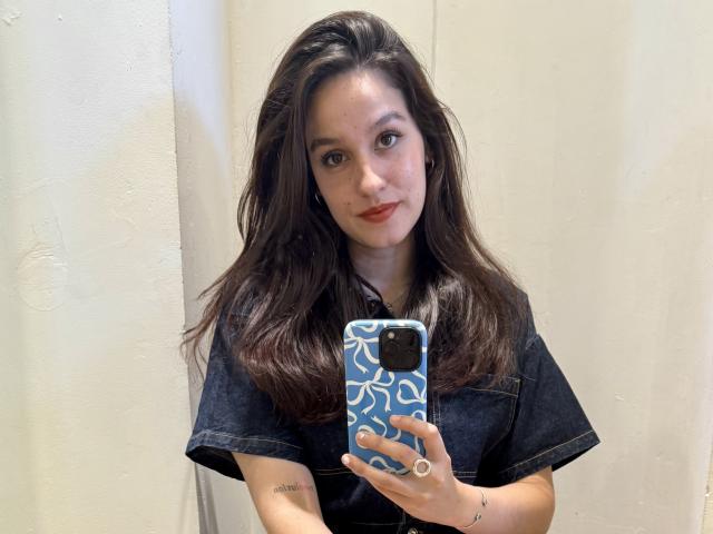 AnastasiaXO, con cabello castaño ondulado, vestida con una camisa vaquera, sosteniendo un teléfono azul con patrones blancos, tomando un selfie, mostrando tatuajes en su brazo derecho..