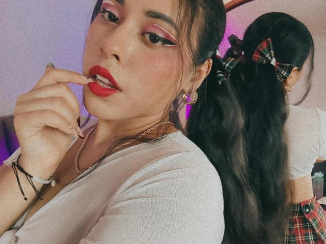 MarilynHills with red lipstick, pink eyeshadow, and finger in mouth, wearing white shirt, black bracelet, and gold necklace, with long black pigtails in plaid bows.