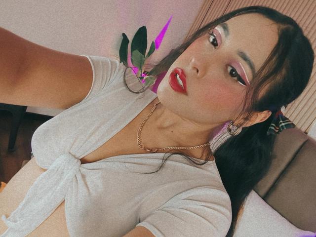 MarilynHills with red lipstick, pink eyeshadow, tied-up white top, showing big tits, gold necklace, black pigtails, sexy face, holding a plant with pink leaves.