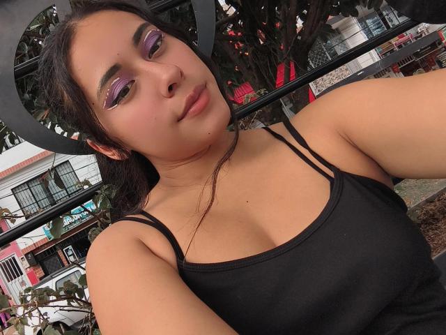 MarilynHills has purple eyeshadow, black tank top with straps, brown hair, and a cute face. Boobs are visible, and she's sitting with a slightly bored expression.