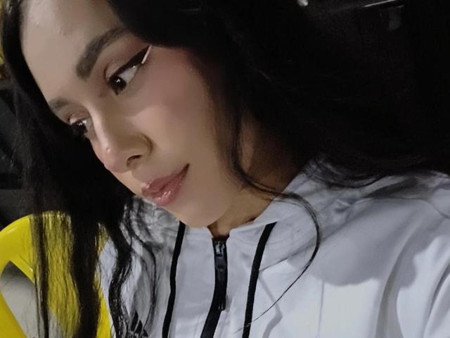 MarilynHills has long black hair, brown eyes, and a slight smile with glossy pink lips. She's wearing a white zip-up hoodie, looking down with a relaxed expression.