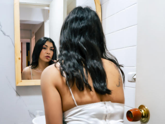 ChanelBrouw with long black hair, back to mirror, looking at reflection, wearing white strap dress, visible tattoo on right shoulder, standing in bathroom, door with brass handle.