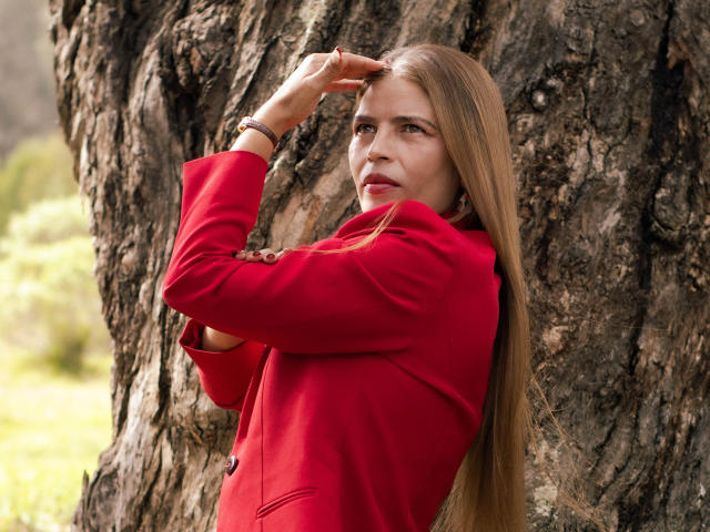 ParisMatur69, avec de longs cheveux blonds, un manteau rouge, appuyée contre un arbre, levant son bras droit, regardant de côté avec un léger sourire, fond vert flou..