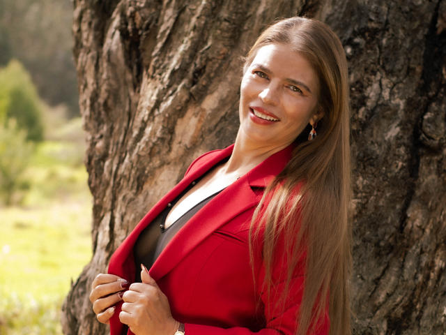 ParisMatur69 con lunghi capelli biondi, un blazer rosso, una maglietta nera e orecchini, in piedi contro un albero, sorridente, che tiene il blazer, mostrando il seno, in un ambiente esterno soleggiato..
