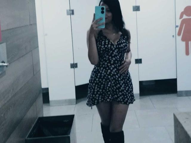 MelodyLauries in a black floral dress with a plunging neckline, holding a phone in front of her face, showing her tits and ass, wearing black knee-high boots.