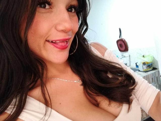 MelodyLauries has dark brown wavy hair, pink lipstick, hoop earrings, and a white top showing her big tits. She's smiling with her tongue out, wearing a silver necklace.