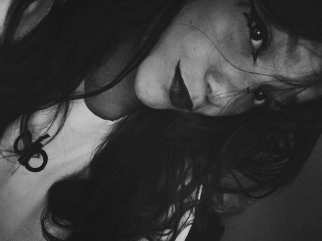 MelodyLauries with dark lipstick and wavy hair, looking up with a slight smile in a black-and-white photo. Her tits are partially visible under a white top.