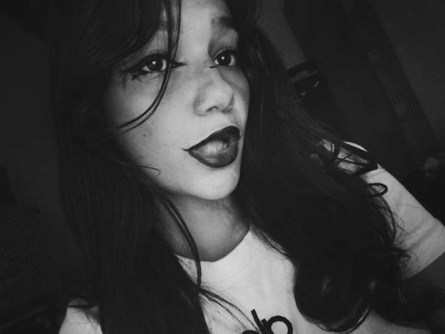 Black-and-white selfie of MelodyLauries with long dark hair, dark lipstick, and tongue out. She's wearing a white shirt, and her freckles are visible. Her eyes are half-open.