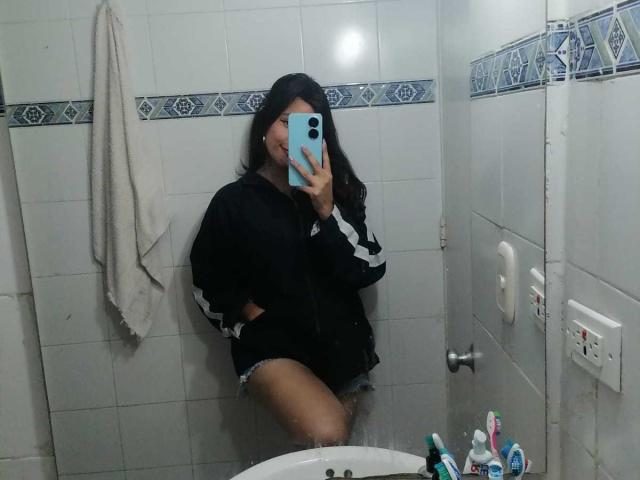 MelodyLauries with long black hair, wearing a black track jacket and shorts, sitting on a toilet, holding a blue phone, showing her ass, in a tiled bathroom.