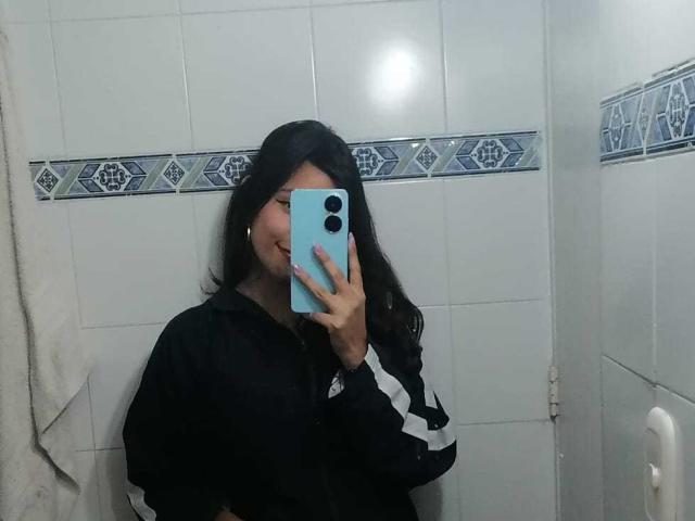 MelodyLauries with long black hair, wearing a black and white shirt, holding a blue phone, covering her face, in a white-tiled bathroom, mirror selfie.
