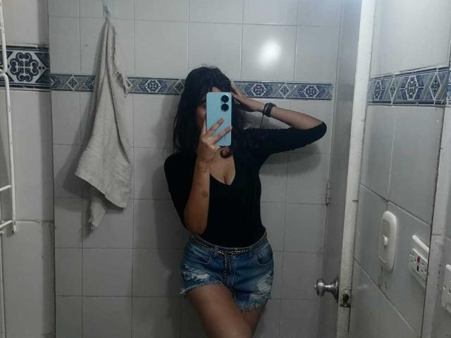 MelodyLauries with medium tits, black top, blue denim shorts, and a silver chain, taking a mirror selfie in a white-tiled bathroom, holding a blue phone.