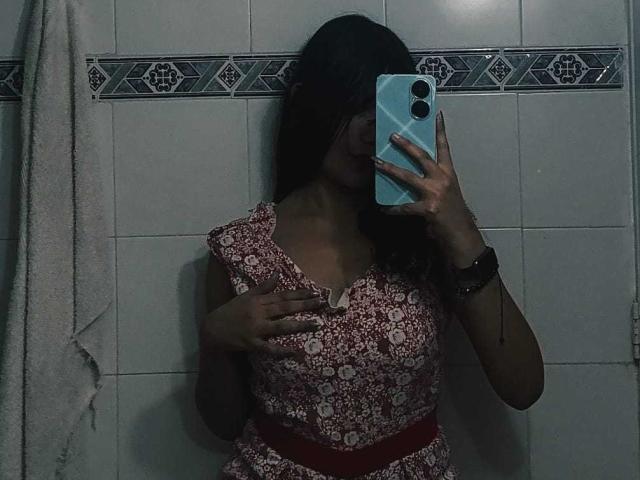 MelodyLauries with dark hair, wearing a red floral dress, holding a blue phone, covering her tits with one hand, standing in a dimly lit bathroom, towel on left.
