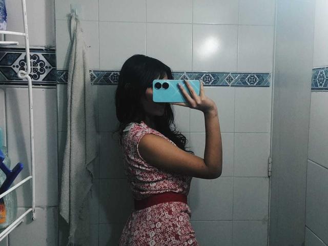 MelodyLauries with long dark hair, wearing a red floral dress, holds a blue phone, covering her face, in a bathroom mirror selfie, showing her side and small ass.