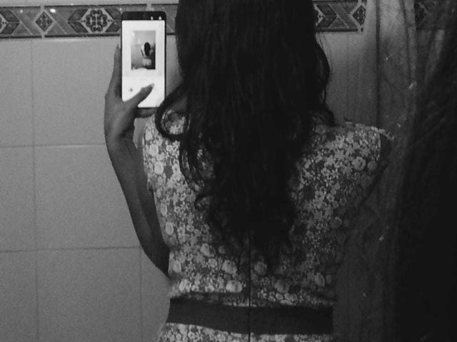 MelodyLauries with wavy dark hair, wearing a floral dress with a black belt, taking a selfie with her phone, showing her back and upper body in a black-and-white photo.