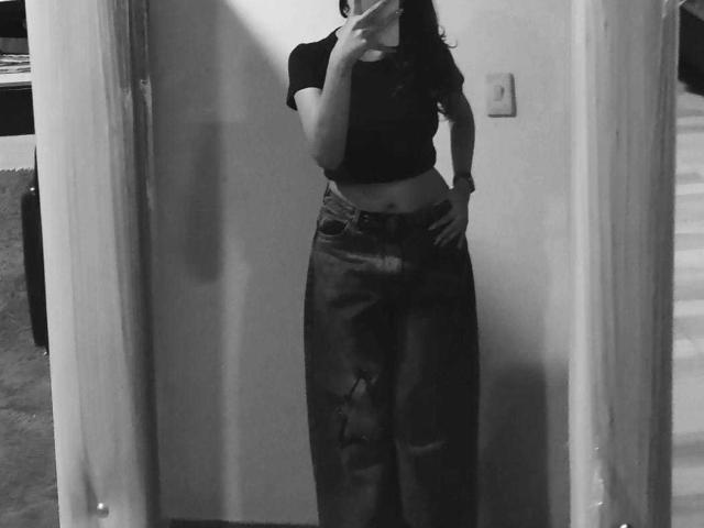 MelodyLauries, black crop top, dark jeans, hand on hip, mirror selfie, black-and-white photo, visible belly button, phone in hand, wavy dark hair, partial face obscured.