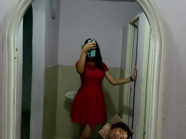 MelodyLauries in a red dress, standing in front of a mirror, holding a phone to cover her face, with her ass slightly visible, in a dimly lit room.