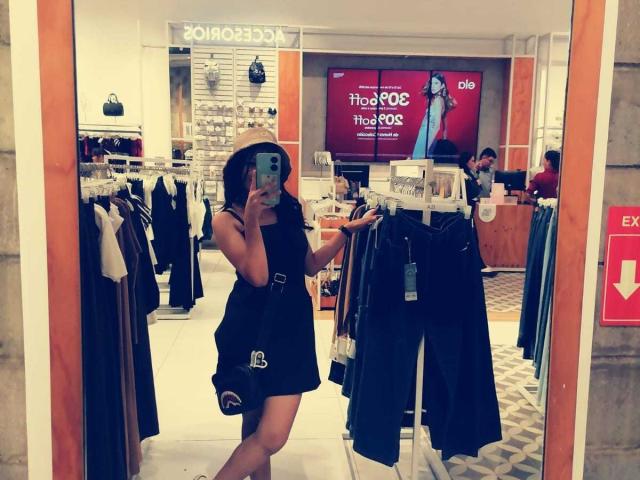 MelodyLauries, with a slim, toned body, wearing a black dress and sun hat, takes a mirror selfie in a clothing store, showing off her ass in the reflection.
