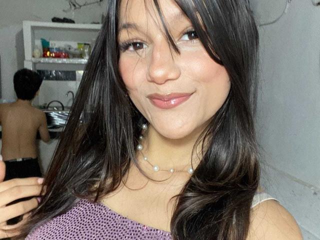 MelodyLauries with long dark hair, shiny lips, and a pearl necklace, smiling at the camera, wearing a purple patterned top, showing her tits. A shirtless guy is in the background.