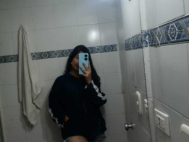 MelodyLauries, wearing a black hoodie and denim shorts, takes a mirror selfie in a tiled bathroom, covering her face with a blue phone. Her hand is in her hoodie's pocket.