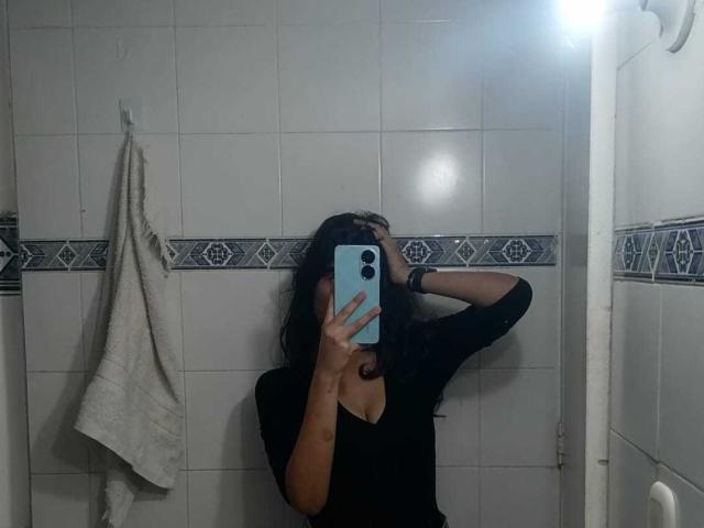 MelodyLauries with long dark hair, black top, and visible tits, taking a mirror selfie in a tiled bathroom, holding a blue phone, hand in hair.