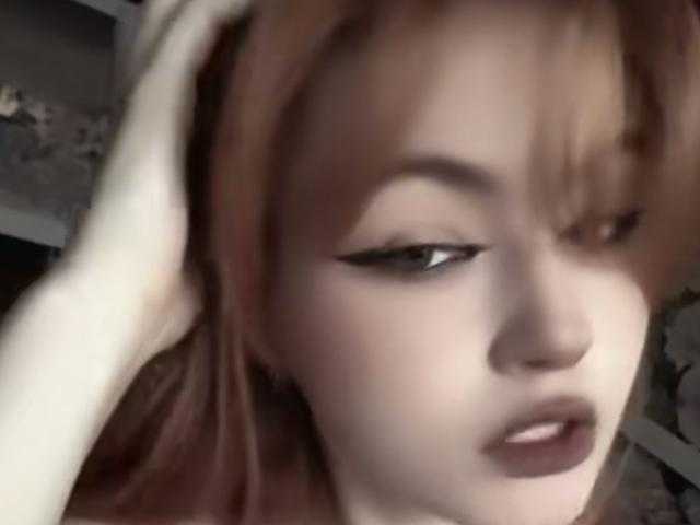 MargSeyler a la peau pâle, les cheveux roux, et elle touche sa tête avec sa main droite. Son eye-liner noir est visible, et ses lèvres sont légèrement entrouvertes..