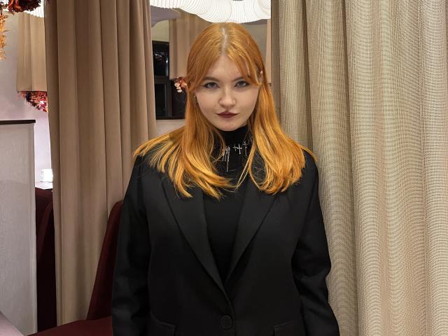 MargSeyler, avec ses cheveux orange, son costume noir et son rouge à lèvres foncé, se tient devant des rideaux beige, avec une expression sérieuse et un collier en forme de croix..