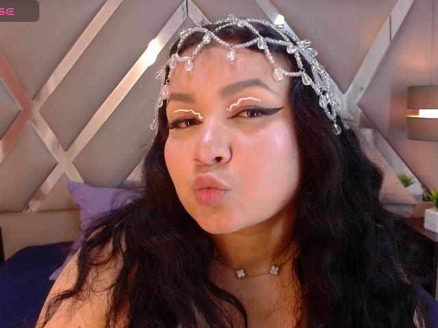 LillyMayer has curly black hair, a silver headpiece with dangling crystals, and is pouting her full lips. She has bold eyeliner and a necklace with small gems.