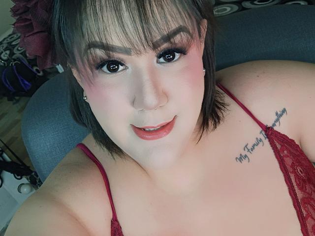 GiiannaMorrys, com cabelo castanho curto, batom vermelho e maquiagem rosa nos olhos, vestindo um sutiã de renda vermelho, mostrando seus seios e uma tatuagem no ombro direito que diz "Minha Família T"..".