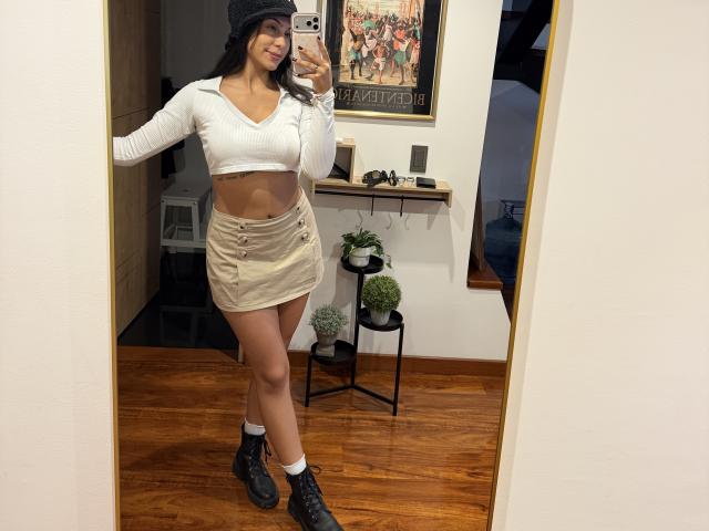 SarahPrincess with tan skin, long dark hair, wearing a white crop top, beige button-up skirt, black boots, and a beanie, showing her small tits and slim ass in a mirror.