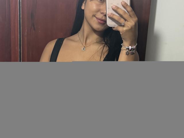 SarahPrincess with large tits in a black tank top, taking a mirror selfie with her phone, wearing a bracelet and necklace, smiling slightly. Brown wood door in background.