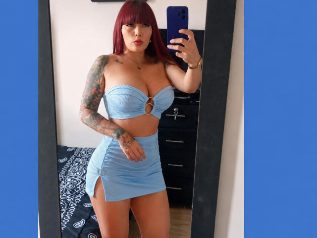 IrisGoddesss with red hair, tattoos, blue strapless top with cutout, blue high-waisted skirt, holding phone, mirror selfie, medium tits, small ass, taking a sexy photo.
