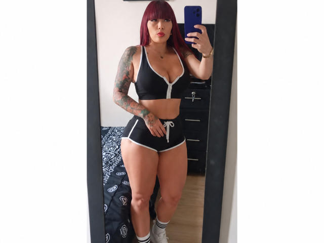 Selfie no espelho de IrisGoddesss com cabelo ruivo, tatuagens no braço direito, top esportivo preto e shorts, meias brancas, segurando um telefone azul, mostrando seu corpo curvilíneo e cheio..