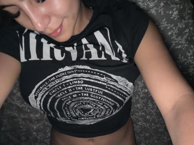 SarahPrincess with medium tits in a black, short-sleeve, graphic t-shirt, lying down, looking at the camera, smiling slightly, dark hair, light skin, subtle makeup, floral-patterned background.