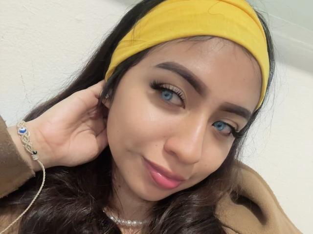 Zoe, con una fascia gialla, occhi marroni e rossetto rosa, sorride leggermente. Ha lunghi capelli scuri, un cappotto marrone e un braccialetto con pietre blu e bianche..