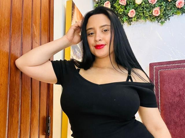 GraceBlue69 con cabello negro largo, lápiz labial rojo, blusa negra con un hombro descubierto que muestra un busto grande, brazo derecho levantado, sonriendo a la cámara, de pie junto a una puerta de madera, decoración floral en la pared..