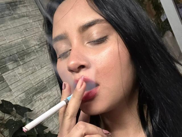 GraceBlue69, avec ses cheveux noirs, les yeux fermés, tenant une cigarette entre ses lèvres rosées, son doigt touchant sa lèvre supérieure. Elle a une peau claire et porte du vernis à ongles rose..