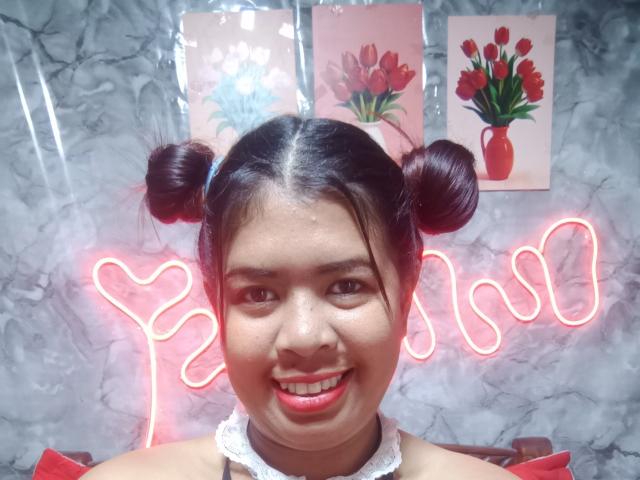MariahSquirting69 with double buns, smiling, red lipstick, white lace collar, neon heart sign, gray marble wall, red tulip paintings, red vase, and red neon lights in the background.