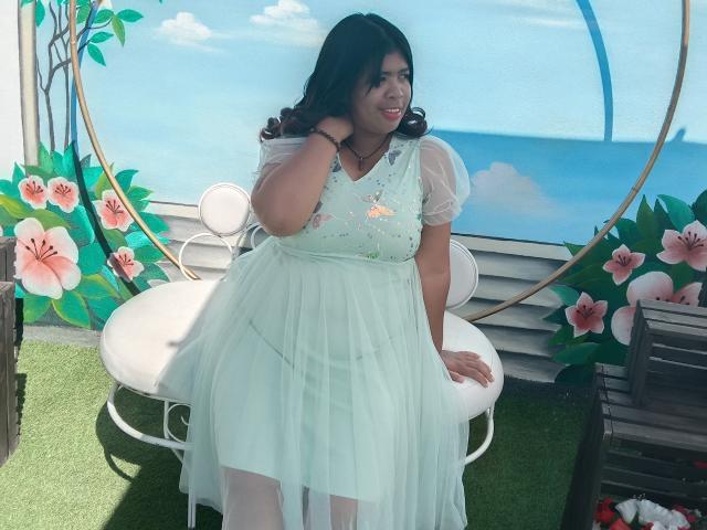 MariahSquirting69 with curvy body, medium tits, and light green dress, sitting on white chair, smiling, hand on head, black hair, sheer sleeves, floral embroidery, green grass background.