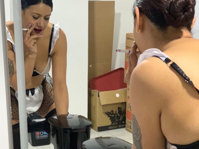 AriannaSeete with dark hair in a bun, wearing a black and white lace bra, polka dot apron, and black strap, smoking in front of a mirror, showing her tits.