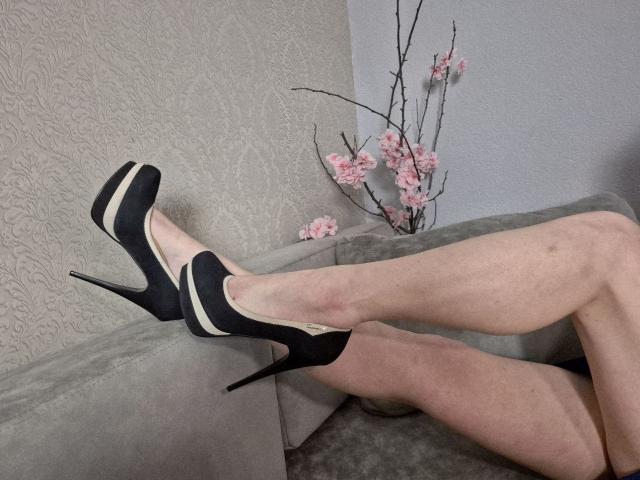 WenTinas Beine liegen auf einem grauen Sofa, sie trägt schwarze High Heels mit weißen Streifen, wodurch ihre helle Haut und eine leichte Bräune sichtbar werden. Im Hintergrund ist ein Zweig mit Blüten zu sehen..