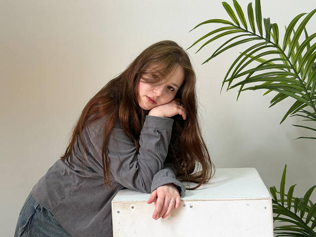 Alica James mit langen braunen Haaren, einem grauen Pullover und blauen Jeans lehnt sich an einen weißen Würfel, ihre Kinn auf ihrer Hand, und blickt mit einem leichten Lächeln in die Kamera..